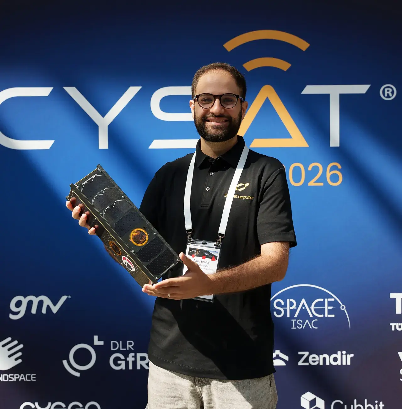 Team member holding satellite hardware in front of the CYSAT sponsor wall